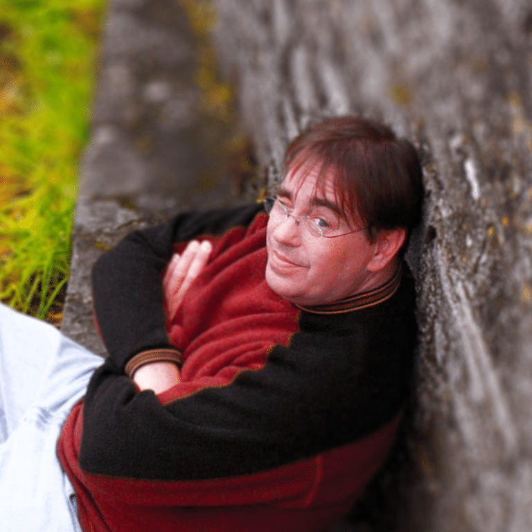 The writer Patrick O'Sullivan, arms crossed, seated and leaning against a stone wall outdoors.